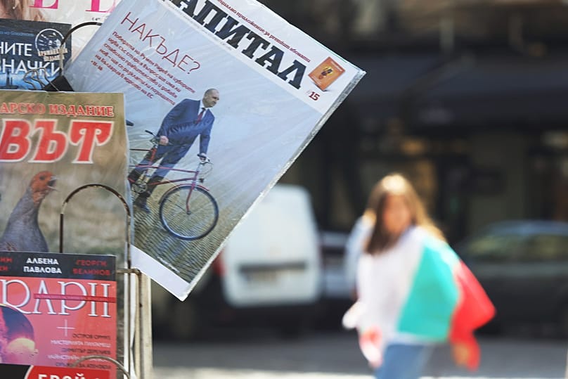A person carrying Bulgarian Flag passes behind newspaper's cover showing the Former President Rumen Radev in Sofia, 20 April, 2026