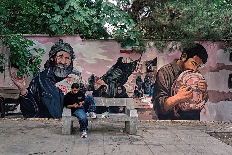 A man on his smartphone in front of a mural honouring children killed in a strike in the southern city of Minab in a suspected US airstrike, in northern Tehran, 15 June 2026