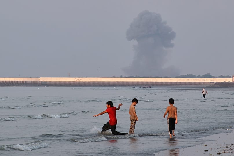 Three boys play in the shallow waters of the Strait of Hormuz, as a plume of smoke rises from an explosion in the background, off Bandar Abbas, Iran, Monday, July 13, 2026. 