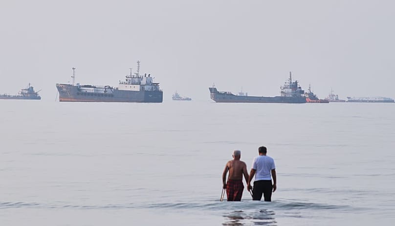 Two men wade in the waters of the Strait of Hormuz with vessels anchored in the background, off Bandar Abbas, Iran, Sunday, July 12, 2026. (Razieh Poudat/ISNA via AP)