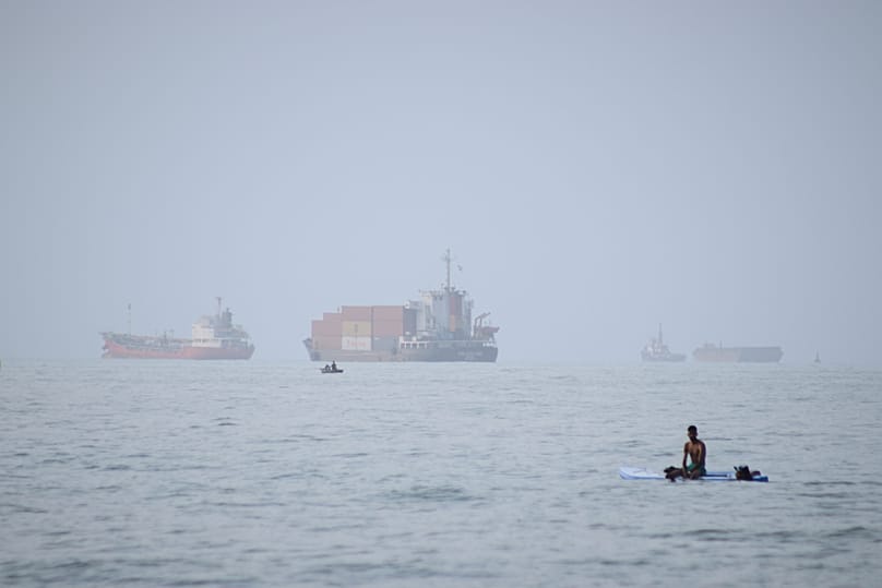 A man sits on a surfboard as a container ship and other commercial vessels appear anchored in the Strait of Hormuz off Bandar Abbas, Iran, Monday, July 27, 2026.