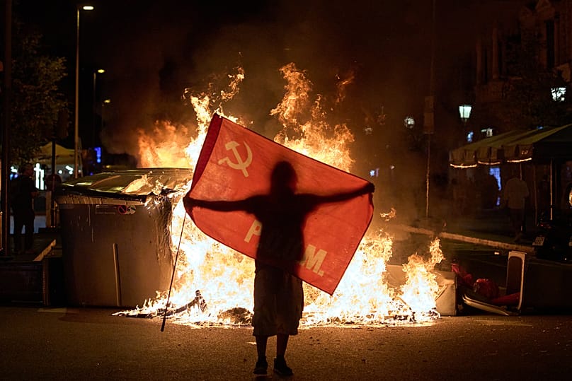A protesters holds a USSR flag next to trash cans on fire during clashes with police in a demonstration over Spain's housing crisis, in Barcelona, Spain, Monday, Oct. 5, 2026