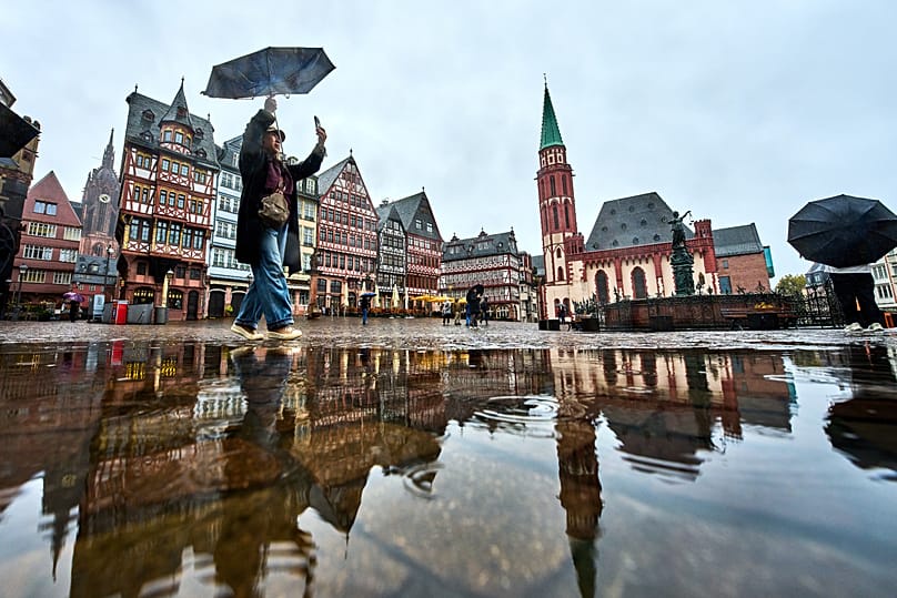 A tourist takes pictures on the Roemerberg square in Frankfurt, Germany, during rain falls on Sunday, Oct. 25, 2026. (AP Photo/Michael Probst)