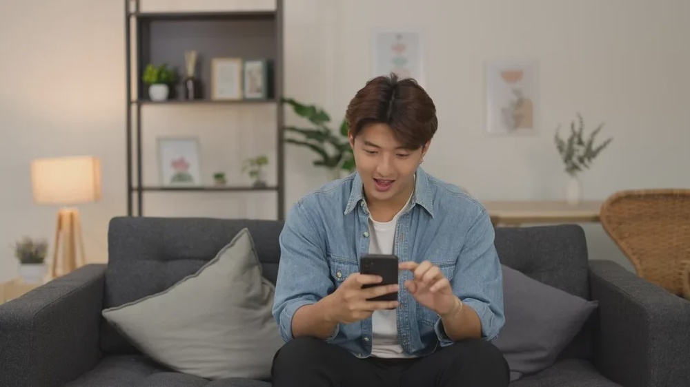 young asian man smiling while using smartphone living room sofa 1308 188892