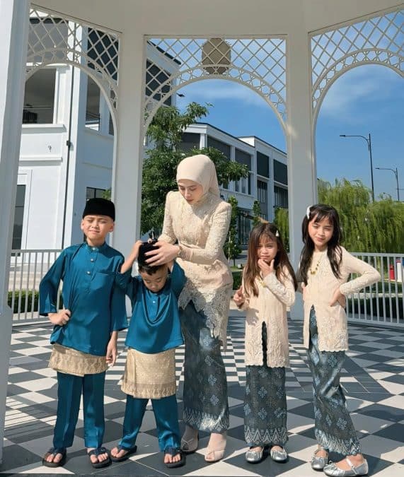 Mother in a beige lace outfit and hijab helps two young boys in blue baju melayu while two girls in cream kebaya smile nearby on a black-and-white checkered patio under white decorative arches.