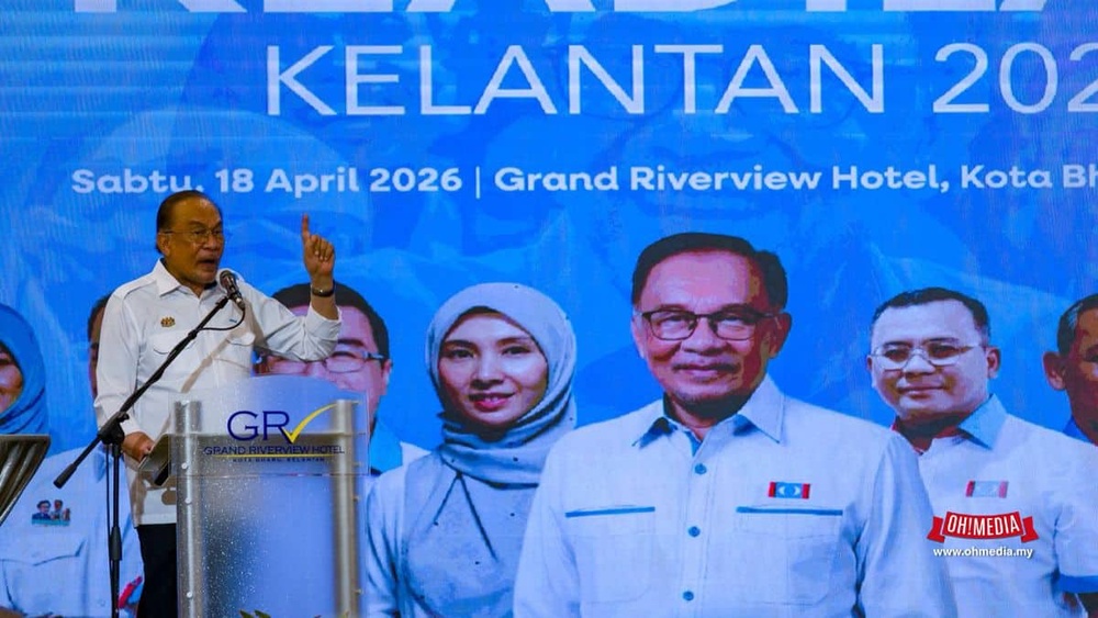 Man in a white shirt speaks at a podium during a public event on a blue backdrop that reads Kelantan 2026, with several officials standing behind him.