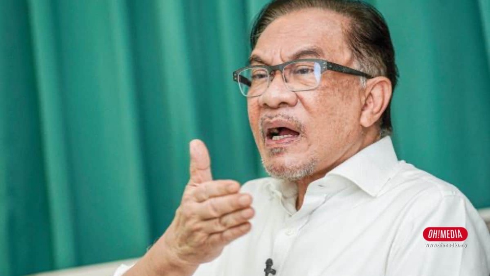 Man with glasses in a white shirt speaks, gesturing with his right hand and wearing a lapel mic.