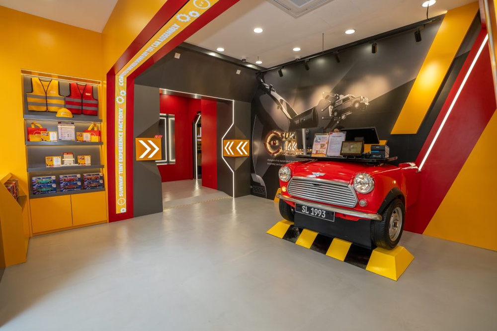 Bright automotive showroom with a red vintage car on a yellow-and-black ramp, yellow walls, and bold graphic wall art on the right.
