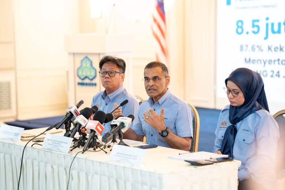 Three officials in blue shirts at a press briefing, speaking into multiple microphones on a table with a large screen and flag in the background.