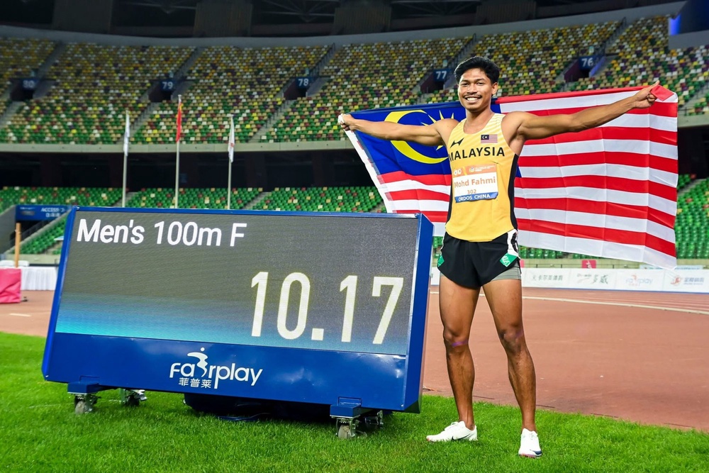 Azeem Fahmi holding the Malaysian flag beside a men’s 100m final results board