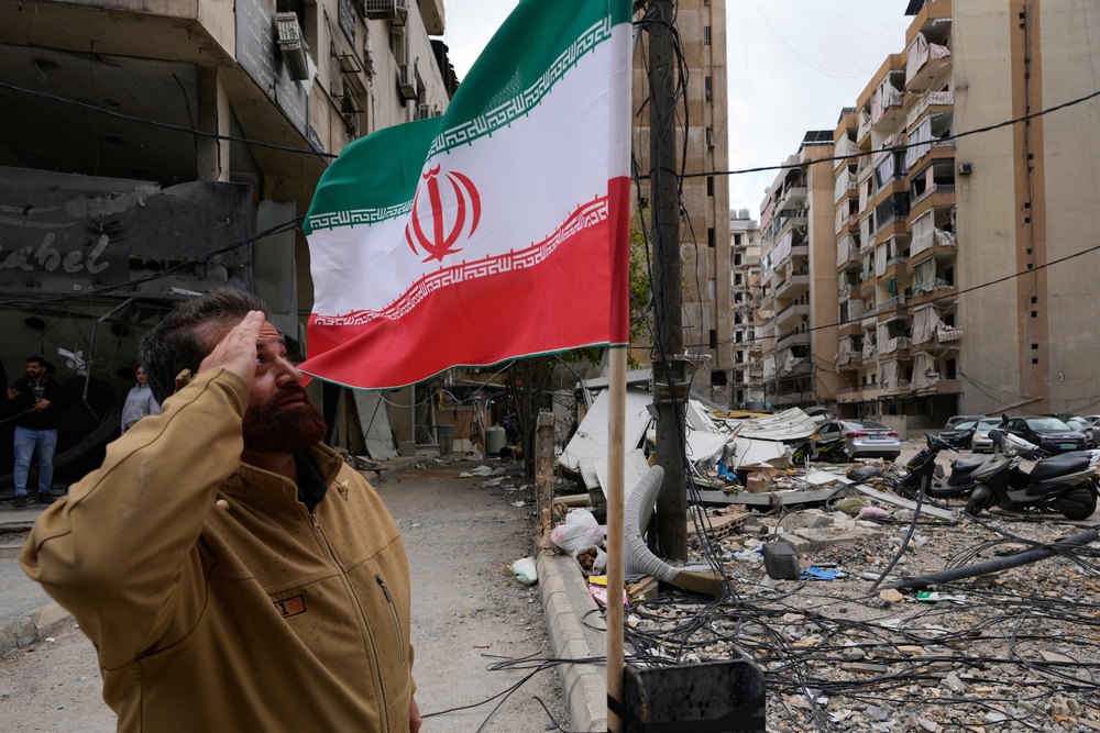 A Hezbollah supporter salutes the Iranian flag in front of a damaged building following a ceasefire between Hezbollah and Israel, in Beirut’s southern suburbs (Hussein Malla/AP) (AP)
