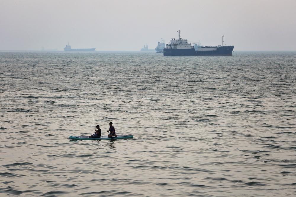 Two boys paddleboard in the sea as ships are anchored near the shoreline on 22 April 2026 in Bandar Abbas, Iran (Getty)