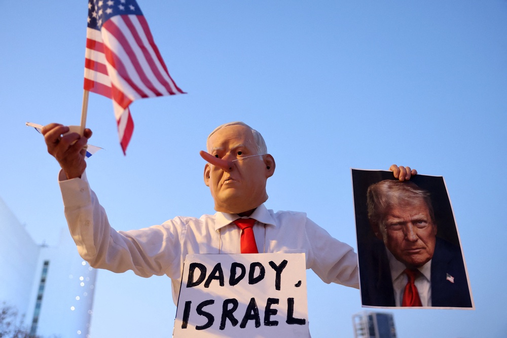 An Israeli left-wing activist, disguised as the Israeli Prime Minister Benjamin Netanyahu and holding an image of the US President Donald Trump and the US flag, demonstrates in HaBima Square against the ongoing war with Iran and against the Israeli government, in Tel Aviv (AFP/Getty)