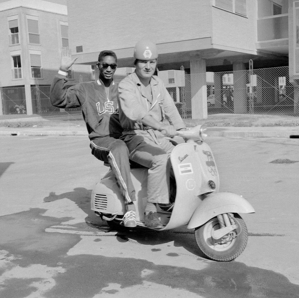 FILE - U.S. high jumper John Thomas, of Cambridge, Mass., waves from a Vespa scooter with an unidentified official Olympic driver inside the Olympic Village, in Rome, Aug. 27, 1960. (AP Photo/Mike Stern, File) (1960 AP)