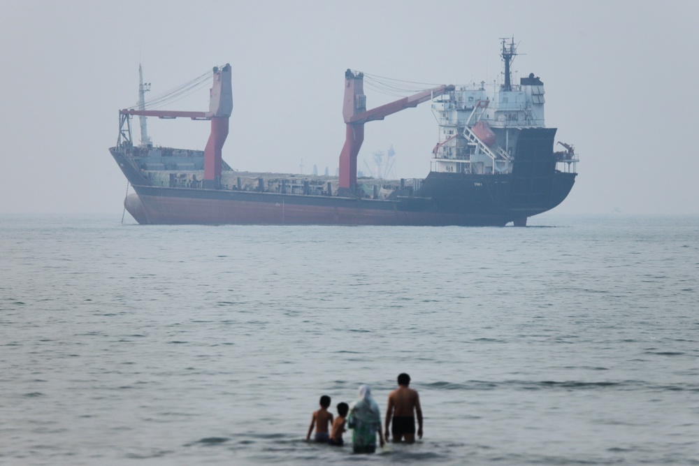 A group of people stand in shallow water as a cargo ship appears anchored in the Strait of Hormuz off Bandar Abbas (AP)