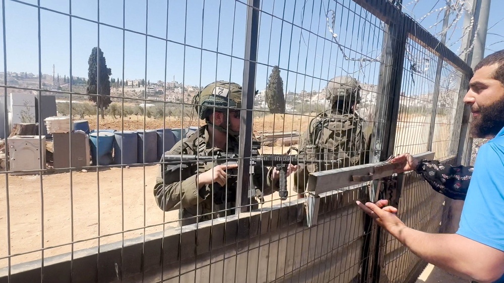 People wearing a military uniform stand behind a fence as a Palestinian man speaks to them from inside his home, during an Israeli settlers' attack on a Palestinian property in the village of Al-Mughayyir, near Ramallah, in the Israeli-occupied West Bank, July 11, 2026 (Reuters)