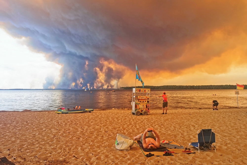 A beach goer looks at his smartphone as a vendor waits with his cart on the beach of Moutchic on the shore of Lake Lacanau (Etang de Lacanau) in Lacanau , southwestern France on July 24, 2026, as clouds of smoke rise into the sky from the Gironde wildfire. (AFP/Getty)