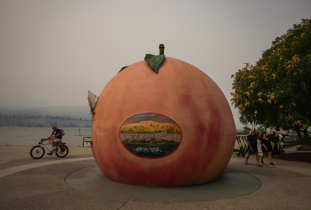 People walk and cycle past an ice cream stand in the shape of a peach as smoke from the Bald Range wildfire envelops Okanagan Lake, in Penticton, British Columbia, on Sunday, Aug. 9, 2026 (AP)