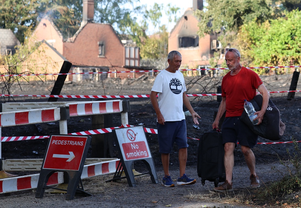 A person carries belongings in a suitcase, and a dog bed under their arm, as they walk near fire-damaged houses after a series of wildfires, in Stourbridge (AFP/Getty)