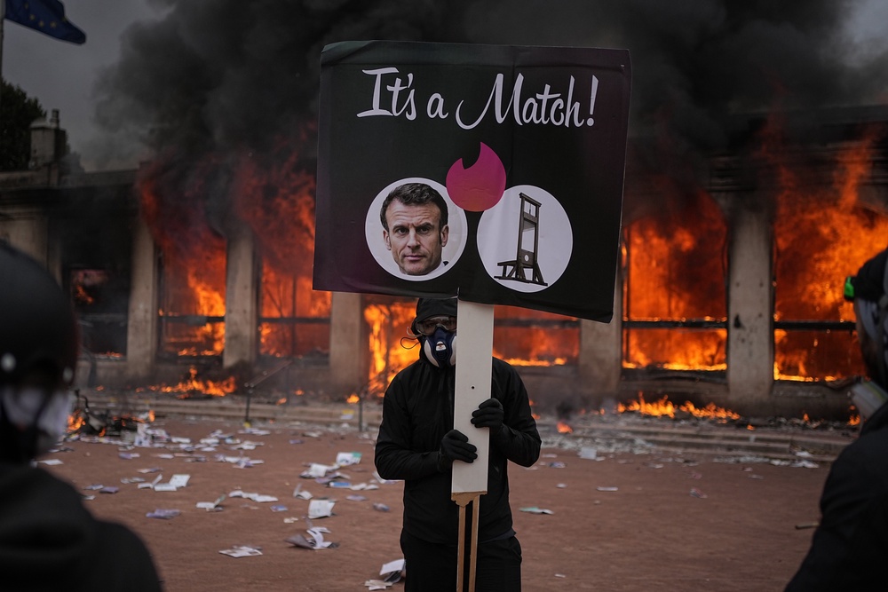 A protester holds a placard with a photo of French president Emmanuel Macron and a guillotine in front of a burning tourism office during a demonstration against rising living costs and in solidarity with students' grievances in Lyon (Copyright 2026 The Associated Press. All rights reserved)