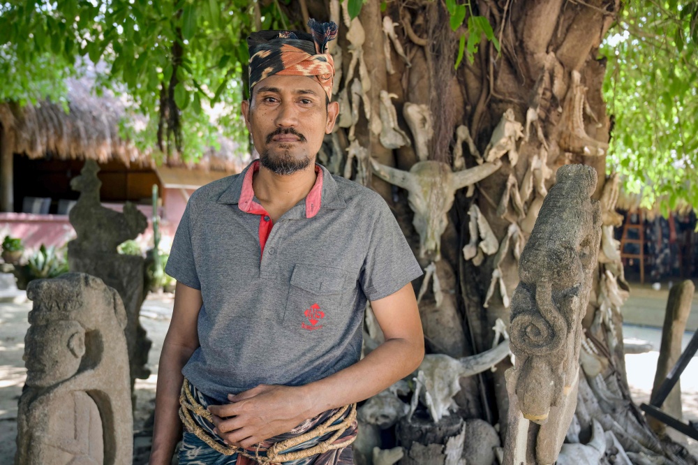 This photo taken on June 24, 2026 shows marapu priest Umbu Remi Deta, who is a maramba (nobleman), posing by a large tree mounted with skulls and jawbones of sacrificial animals alongside traditional stone carvings at Kampung Raja Prailiu at Waingapu in East Sumba, East Nusa Tenggara.