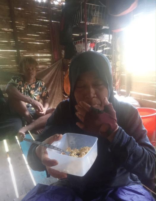 Person in a hoodie and gloves eating cereal from a white plastic container with a spoon inside a wooden shed, another person sitting in the background.