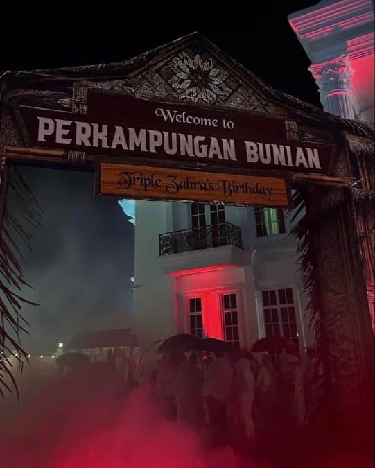 Night scene at a tropical venue with an arch reading 'Welcome to PERKAMPUNGAN BUNIAN' and a 'Triple Zahra's Birthday' sign, red fog and a crowd.