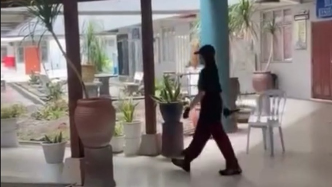 Person wearing a hat and dark clothing walks through a sunlit courtyard with potted plants and white plastic chairs.
