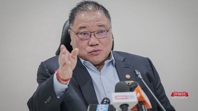 Man in a dark suit and light-blue shirt speaks at a press conference, gesturing with his right hand in front of microphones.