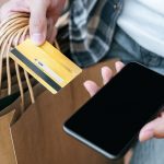 Person shopping with credit card and smartphone while carrying retail bags, representing cashless consumer spending
